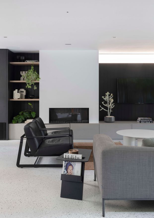 Contemporary living room with a restrained black, white and grey palette. The room features a black built-in shelving unit with plants and decorative objects, a linear fireplace set into pale cabinetry, a dark vertical-panel TV wall, recessed ceiling lighting, black leather lounge chairs, a grey upholstered sofa and a round white coffee table. The floor is a light speckled hard surface, creating a clean, refined interior.