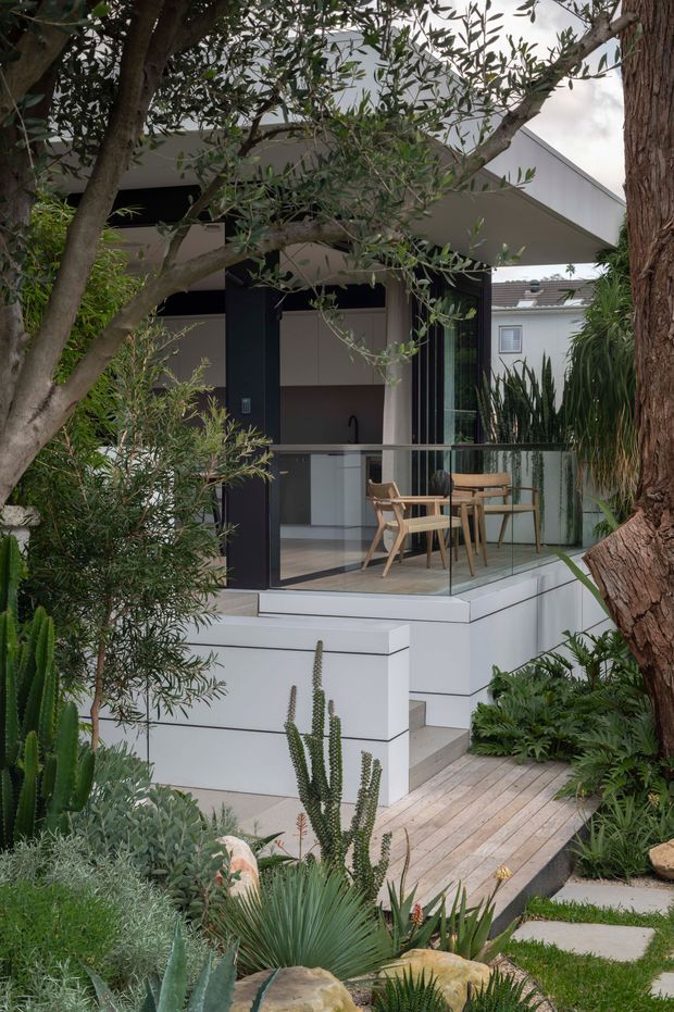 Exterior view of a contemporary compact home extension or pavilion, partially screened by mature trees and dense drought-tolerant planting. The elevated white-painted structure has a sharply angled roof, dark-framed full-height glazing, and a glass balustrade around a small raised terrace. A timber staircase and boardwalk lead through the landscaped garden, while a simple dining setting is visible inside or on the terrace.