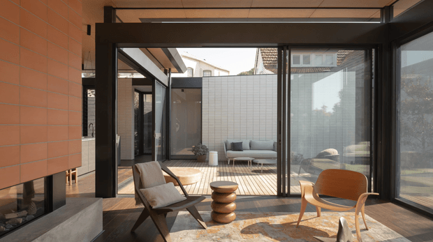 A refined contemporary living room with extensive black-framed glazing opening onto a private courtyard. The room features a suspended terracotta-toned tiled fireplace wall, dark timber flooring, a neutral sofa, sculptural timber side table, mid-century-inspired lounge chairs, and a large patterned rug. The glazed courtyard and adjacent tiled wall create a bright indoor-outdoor connection.
