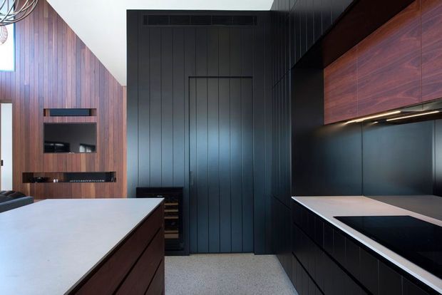 A sleek contemporary kitchen with dark vertical wall panelling, rich timber cabinetry, black lower joinery, and a large pale island benchtop. Integrated appliances and under-cabinet lighting create a refined, high-contrast interior, with a media niche visible on the adjacent timber-lined wall.