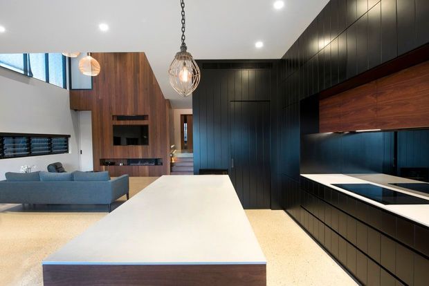 Contemporary open-plan kitchen and living area with a large white island, dark vertically panelled cabinetry, warm timber joinery, integrated appliances, and polished concrete-style flooring. A timber feature wall and lounge area are visible beyond the kitchen, with sculptural pendant lighting and recessed ceiling lights.