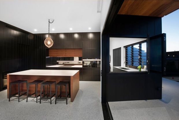 Modern open-plan kitchen with a large timber island and four stools, dark vertically grooved cabinetry and walls, a warm timber ceiling, integrated appliances, and a sculptural pendant light. The right side shows a large opening or servery connecting to an outdoor area, with blue-toned cabinetry and external louvres visible beyond.