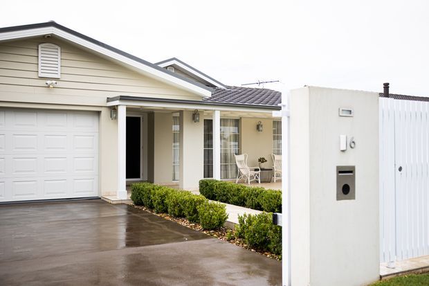 Street-facing view of a single-storey suburban home with a pale horizontal weatherboard facade, gabled roof, attached garage, covered front entry, white-framed glazing, clipped hedges, and a modern white rendered mailbox pillar. The driveway is wet, and the house presents a neat, restrained traditional coastal-suburban appearance.