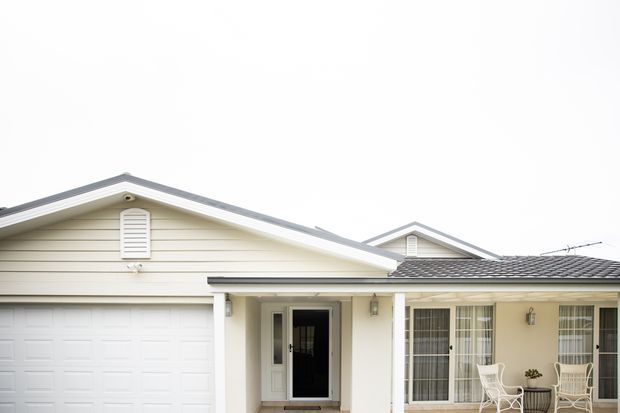 Front exterior view of a single-storey suburban home with cream horizontal weatherboard cladding, a gabled roof, attached double garage, covered entry porch, and large glazed windows with an outdoor seating area.