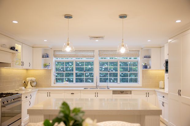 Bright, spacious traditional-style kitchen with extensive cream-white cabinetry, a large central island, pale tiled splashback, integrated appliances, pendant lights, and two wide windows overlooking greenery. The room has a warm, inviting finish with decorative open shelving and classic hardware.