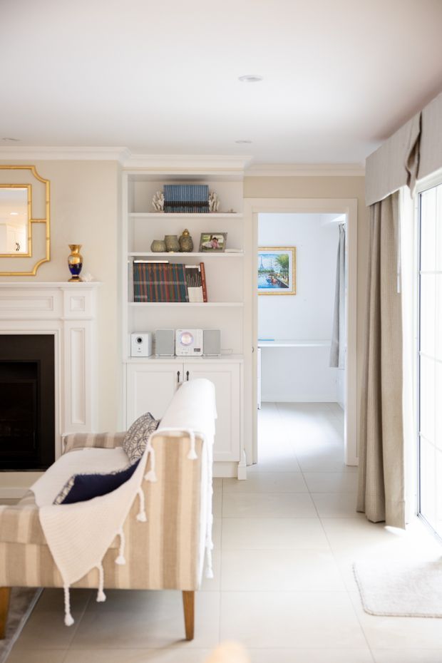 Bright, refined living room with a white traditional fireplace, built-in shelving and cabinetry, decorative books and objects, a neutral upholstered sofa, and tall curtained glazing. A tiled passage leads toward a secondary room with framed artwork visible beyond.
