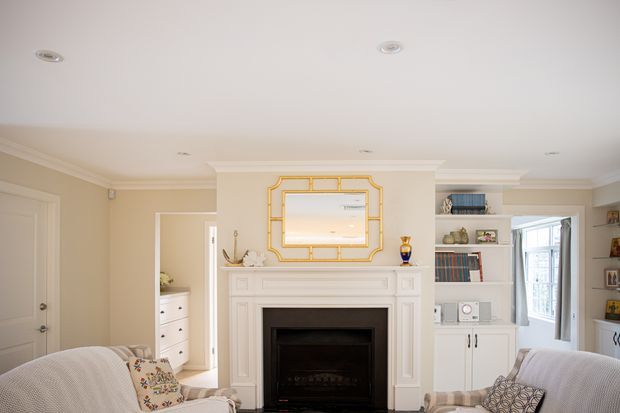Bright, traditionally styled living room with a white detailed fireplace mantel and black firebox as the focal point. Built-in white cabinetry and open bookshelves flank the fireplace, while a large gold-trimmed rectangular mirror hangs above it. Pale upholstered sofas with patterned cushions frame the foreground, and a glazed doorway opens to another light-filled area.