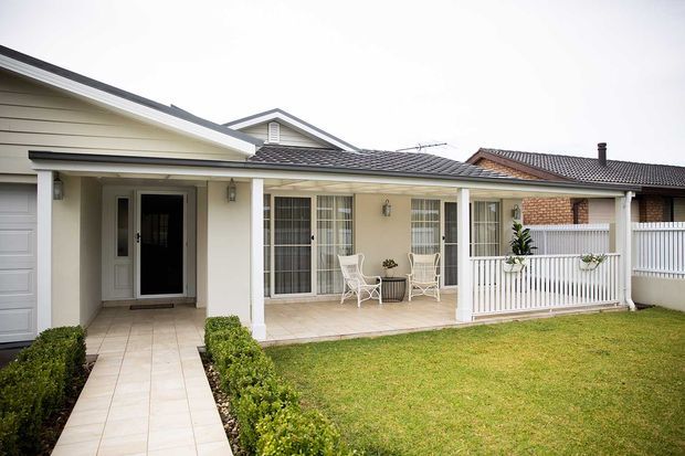 Single-storey suburban house exterior with a covered front veranda, pale rendered walls, tiled pitched roof, white posts and balustrade, glazed entry doors, paved walkway, and a neatly landscaped front lawn.