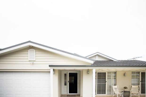 Front exterior view of a single-storey suburban house with a pale horizontal weatherboard facade, prominent gabled roofline, attached double garage, covered entry porch, and a tiled-roof extension with glazed windows and a small outdoor seating area.