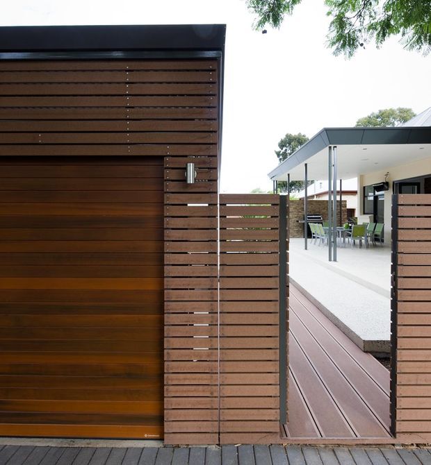 Exterior architectural photograph showing a contemporary residential entrance and courtyard connection. A dark stained timber-clad gate and horizontal timber privacy screen dominate the foreground, opening toward a timber deck and covered outdoor dining or entertaining area. The building has a crisp flat roof, white soffit and walls, dark metal fascia, slim posts, and clean-lined modern detailing.