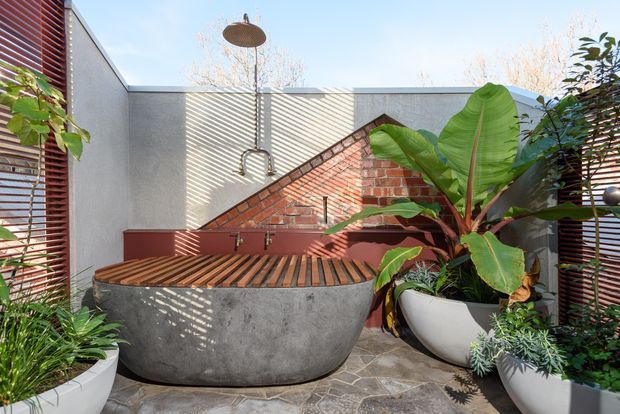A compact outdoor bathroom courtyard featuring a freestanding concrete soaking tub, exposed red-brick wall, open rain shower, timber privacy screens, large tropical plants, and irregular stone paving. The space is bright and lush, with a strong blend of raw masonry and contemporary outdoor bathing elements.