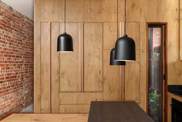 Interior view of a contemporary kitchen or dining area featuring a full-height timber panelled wall with integrated cabinetry, exposed red brickwork, and two large matte-black pendant lights. A dark island or table occupies the foreground, while a narrow glazed door or window reveals brickwork and greenery beyond. The material palette is warm timber, red brick, black metal, and dark wood.