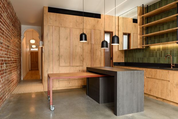A warm, character-filled contemporary kitchen with exposed red brick walls, extensive natural timber panelled cabinetry, a dark island with grey wood-look side panels, and a green vertical-tile splashback. Black pendant lights hang above the island, while a movable timber-topped table or workbench sits in the foreground. An arched hallway and patterned floor tiles add heritage detail, and open timber shelving with books is visible at right.