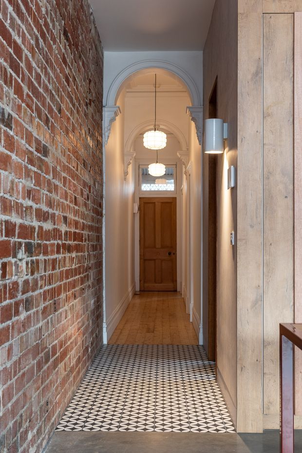 A narrow, refined hallway combining an exposed red brick wall with pale timber wall panelling and warm plasterwork. The entry floor features a bold black-and-white geometric patterned tile transitioning to timber boards, while a sequence of arched doorways, decorative mouldings, pendant lights and a timber door create depth toward the rear of the corridor.