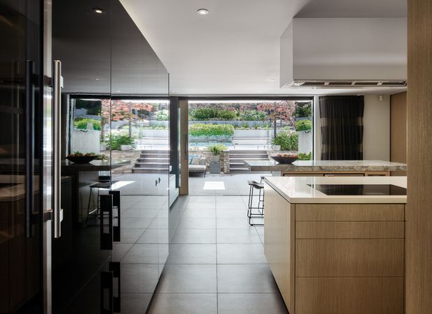 Contemporary open-plan kitchen viewed toward a landscaped stepped courtyard through large glazed openings. The kitchen features a central island with pale stone benchtops, warm timber-grain cabinetry, dark full-height joinery, integrated appliances, recessed ceiling lights, and large-format grey floor tiles. Outdoor planting and terrace steps are visible beyond the glazing.