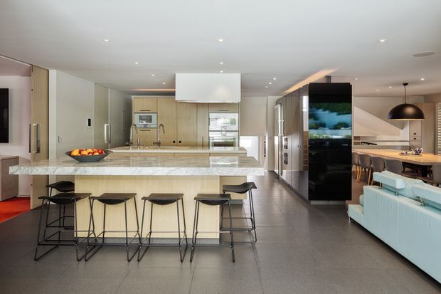 Open-plan contemporary kitchen and living area with a large pale stone island, integrated light timber cabinetry, stainless-steel appliances, dark tiled flooring, and four bar stools. A dining area and pale blue sofa are visible to the right, with a large black-framed glazed or reflective partition and pendant lighting adding contrast.
