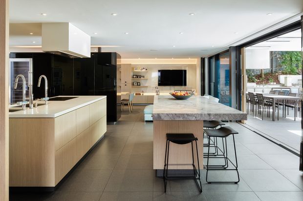 Open-plan contemporary kitchen and living area with a large island, integrated dark cabinetry, pale timber-look joinery, marble-look island benchtop, and bar seating. Floor-to-ceiling sliding glazing opens onto an outdoor dining terrace, while a built-in media wall and dining area are visible beyond the kitchen.