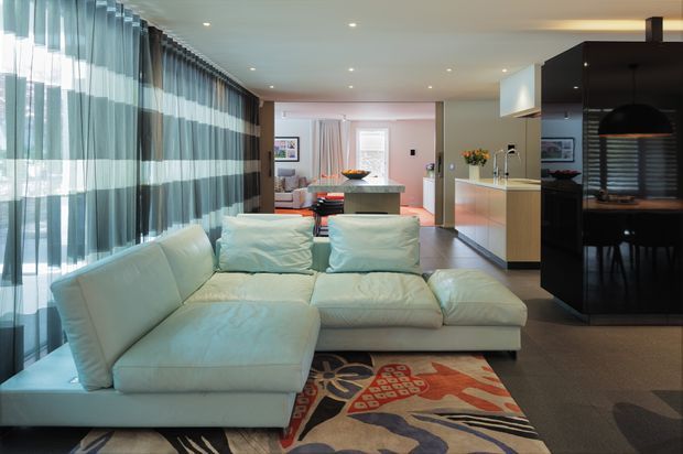 Open-plan contemporary living area with a large pale sectional sofa in the foreground, expansive sheer floor-to-ceiling curtains along the left side, a colourful patterned rug, and a modern kitchen and dining area beyond. The space has recessed ceiling lights, dark kitchen cabinetry, a light island benchtop, and a black pendant light, creating a refined, spacious interior.