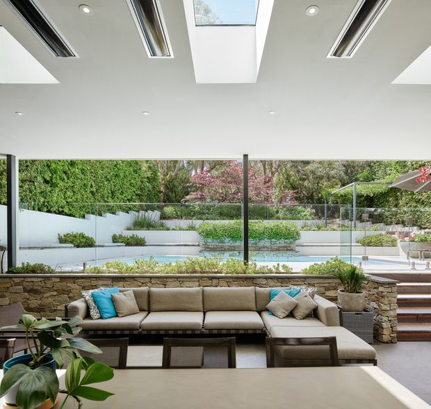Bright contemporary living room opening onto a landscaped outdoor pool area. The room features a large sectional sofa, low coffee table, extensive floor-to-ceiling glazing, a stacked natural-stone feature wall, polished light flooring, and a white ceiling with multiple linear skylights and recessed lights. The garden, pool, and outdoor terrace are clearly visible beyond the glazing.