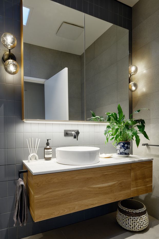 A contemporary bathroom vanity viewed straight on. The room features a floating timber-veneer vanity with a white stone benchtop, round white vessel basin, wall-mounted chrome tapware, large mirrored cabinet or mirror, dark square wall tiles, light grey large-format wall tiles, integrated strip lighting, globe wall lights, and a leafy plant. A towel rail and decorative stool are visible below and beside the vanity.