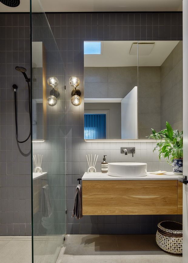 A contemporary bathroom with a walk-in glass shower on the left, charcoal-grey tiled walls, a floating natural timber vanity, white vessel basin, and large frameless mirror. Globe wall lights flank the mirror, while a skylight and pale ceiling brighten the room. A leafy plant and woven basket add warmth and texture.