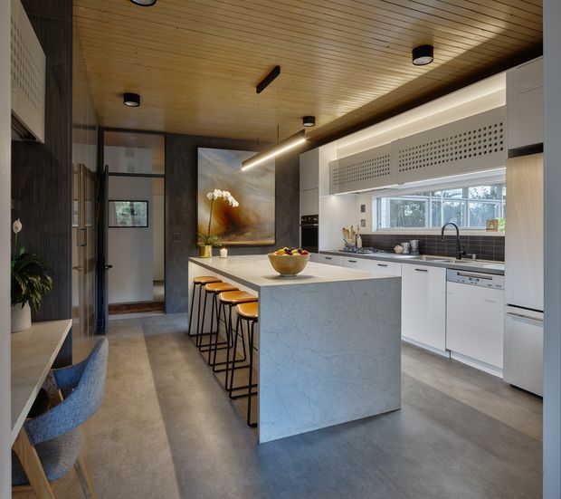 A contemporary open-plan kitchen with a large pale stone island and integrated seating for four. White cabinetry runs along the back wall beneath perforated white overhead panels, with a dark tiled splashback, stainless or dark metal tapware, and a wide window. The room features a warm timber-lined ceiling, polished concrete-look flooring, dark-framed glazing and doors, and a linear pendant light. A dining/work nook with blue upholstered chairs is visible at left, while an artwork and indoor plant add decorative detail.
