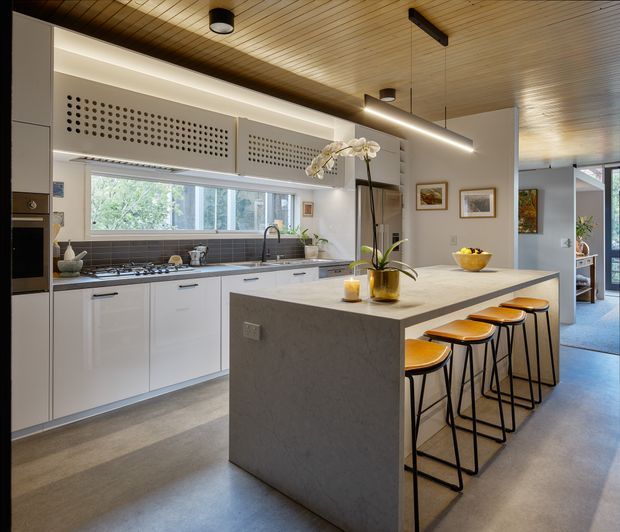 A bright, open-plan contemporary kitchen with a large pale stone island and seating for four. White flat-panel cabinetry lines the rear wall beneath a wide window, with a dark tiled backsplash, integrated appliances, and a long linear pendant light. The kitchen features a warm timber slatted ceiling, polished grey concrete-style flooring, black-framed stools, and subtle decorative styling. A living or circulation area is visible beyond the kitchen at right.