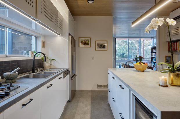 A bright, contemporary galley kitchen viewed along the working aisle toward a dining and living area. White handleless-style cabinetry with dark pulls lines both sides, with a large pale stone island and integrated appliances. The kitchen includes a stainless-steel sink and tap, dark tiled splashback, wide glazing, timber-lined ceiling, integrated linear lighting, open shelving, and indoor plants. The space feels refined, functional, and welcoming.