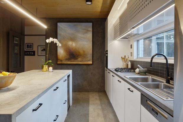 A refined contemporary kitchen featuring a large pale stone island with white cabinetry and black hardware, a matching galley run with stainless-steel double sink and integrated appliances, dark tiled backsplash, warm timber ceiling, and a dark textured feature wall with artwork. Linear pendant lighting and broad windows provide a bright, polished atmosphere.