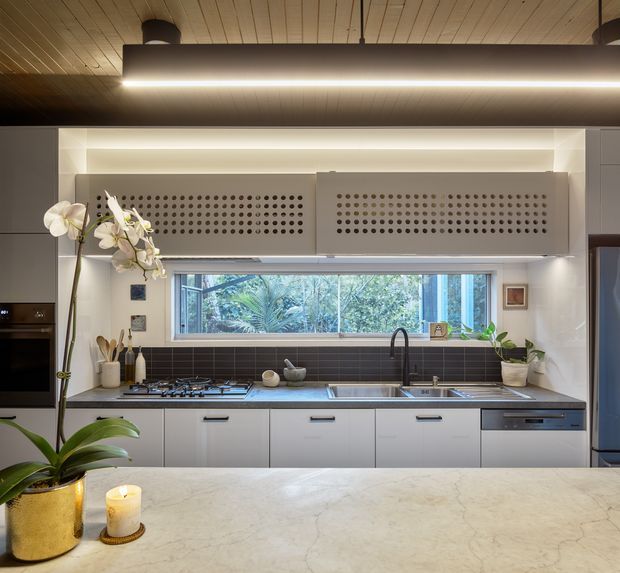 A contemporary kitchen viewed from behind a large pale stone island. The room features a long run of light grey cabinetry, dark horizontal tile splashback, integrated cooktop and oven, stainless-steel sink with dark tapware, and distinctive perforated overhead cabinet doors. A wide garden-facing window brings in natural light, while warm timber slatted ceilings and a linear pendant create a refined, warm atmosphere. Indoor plants and an orchid add organic accents.