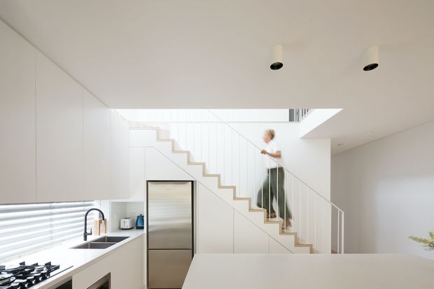 Bright minimalist open-plan kitchen and stair hall with seamless white cabinetry, a pale benchtop, stainless-steel refrigerator, black fixtures, and a sculptural open-riser timber-edged staircase beneath a large double-height void. Horizontal window blinds provide filtered daylight, and the space has a clean, highly restrained contemporary appearance.