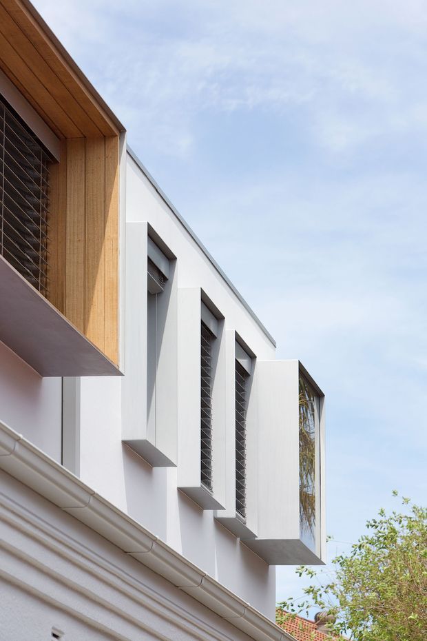 Upward-looking exterior view of a contemporary multi-level building facade. White rendered walls feature projecting vertical window bays with dark louvred or screened glazing, alongside a warm timber-lined projecting volume and a tall glazed opening. A lower section has traditional guttering and cornice-like detailing, creating a modern addition or facade composition against a pale sky.