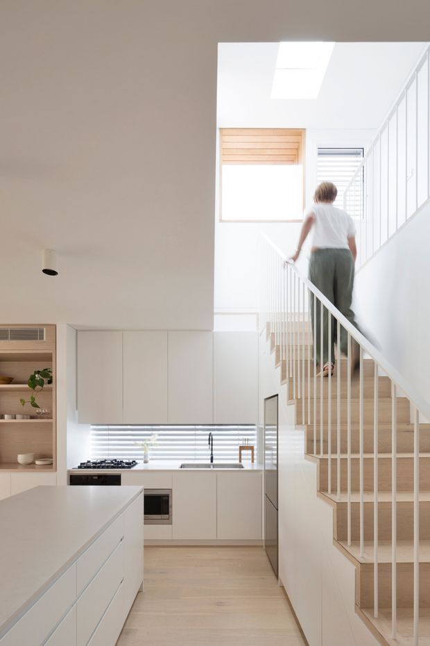 Bright contemporary open-plan kitchen and stairwell interior. The image shows a white kitchen with a large island, integrated cabinetry, stone-look surfaces, timber flooring, and a gas cooktop beneath horizontal blinds. A light timber staircase with slim white vertical balustrades rises beside the kitchen toward a sunlit upper level, with a skylight and clerestory-style glazing contributing to the airy atmosphere.
