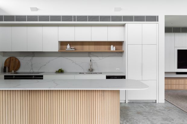 A bright, refined contemporary kitchen dominated by white full-height cabinetry, a long white marble-look stone benchtop and splashback, and a large island with a vertical light timber slatted front. The kitchen includes integrated appliances, a linear timber display niche, open shelving, and a stainless-steel tap. A partial living area with matching timber joinery and a wall-mounted television is visible beyond the opening.