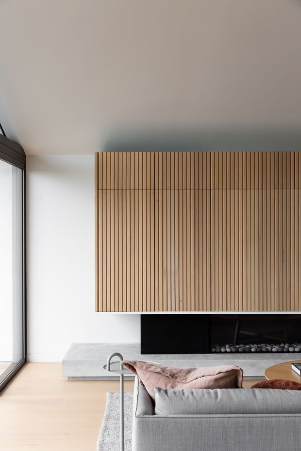 Contemporary living room with a large floor-to-ceiling window, pale timber flooring, a grey sectional sofa, and a long linear fireplace set beneath a prominent vertical timber slatted feature wall. The restrained neutral palette and clean built-in detailing create a refined, warm minimalist interior.