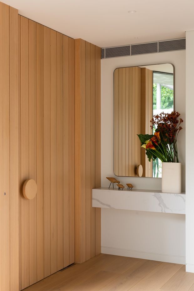 A refined residential entryway featuring full-height vertical timber wall panelling and matching concealed cabinetry, warm timber flooring, and a floating console with a white veined stone benchtop. A rounded rectangular mirror, sculptural small timber objects, and a tall floral arrangement add visual interest. The space has a clean white ceiling with a linear ventilation grille and a calm, warm contemporary aesthetic.
