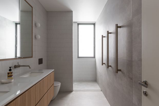 A bright, contemporary bathroom with a long floating timber vanity and white integrated countertop on the left, a large framed mirror, wall-mounted basin tap, concealed toilet, and a walk-in shower at the rear. The shower and side walls are finished in pale grey tiled surfaces, with vertical brushed-metal grab rails on the right wall. A tall window provides natural light, and the space has a clean, restrained material palette.