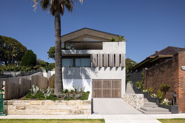 Street-facing view of a contemporary multi-storey residence with a pale rendered facade, broad horizontal glazing, timber-look vertical screening, a flat roof, and an integrated garage. Stone retaining walls, concrete steps, landscaped planting, and a tall palm frame the property in bright daylight.