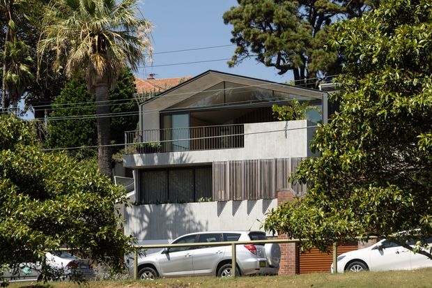 Exterior view of a contemporary multi-level suburban house partially screened by mature trees. The facade combines pale rendered walls, vertical timber-look screening, dark window and door frames, broad balconies, and a prominent pitched gable roof with glazed upper-level frontage. Cars and overhead utility lines are visible in the foreground and surroundings.