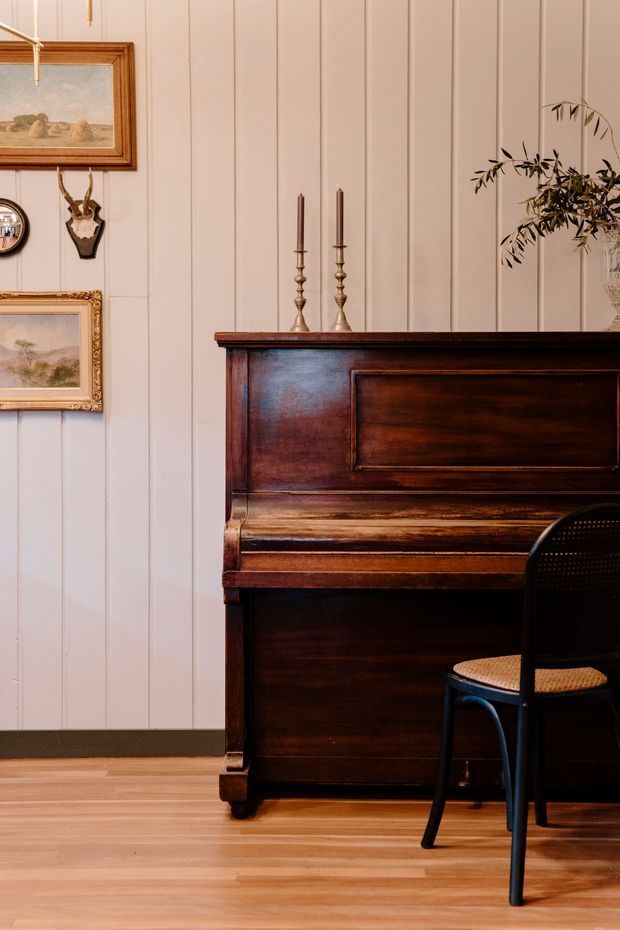 A warmly styled interior vignette featuring a dark antique upright piano against vertical painted wall panelling. Framed landscape artwork, mounted antlers, brass candlesticks, and a leafy branch decorate the wall and piano, while a black bentwood-style chair sits in front. The room has warm timber flooring and a refined, heritage-inspired atmosphere.