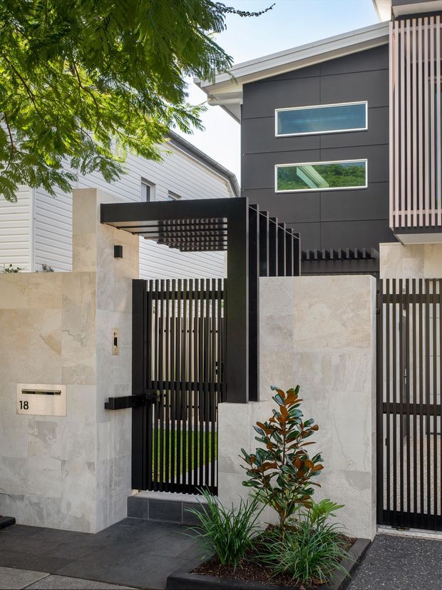 Street-facing view of a contemporary multi-level townhouse facade with dark horizontal cladding, pale stone walls, slim black vertical metal gates, a projecting slatted entry canopy, and narrow horizontal windows. The entry is landscaped with low planting and a tiled forecourt.