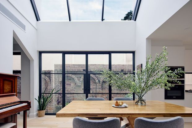 Bright contemporary open-plan dining area with a large natural timber dining table and upholstered chairs, adjacent to a kitchen with dark built-in cabinetry. Full-height black-framed glazing overlooks an exposed brick wall, while a large skylight brings abundant daylight into the double-height space. An upright piano and indoor plants add warmth and character.