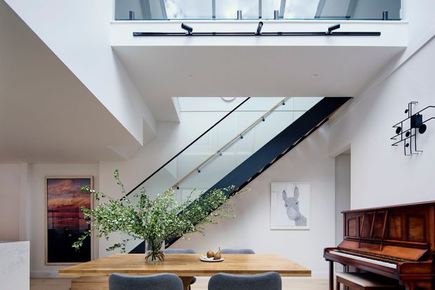 Bright contemporary open-plan interior centred on a timber dining table beneath a dramatic stair void. A black-framed glass balustrade and dark stair structure lead to an upper level, with white walls, a skylight or roof glazing, recessed lighting, artwork, a sculptural wall light, and an upright piano along the right wall. The space feels airy, refined, and architecturally focused.