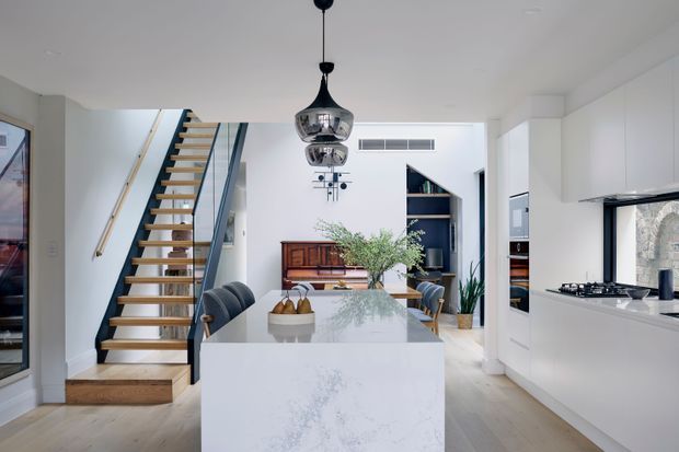 A bright, open-plan contemporary kitchen and dining area with a large white stone island, integrated white cabinetry, a gas cooktop, timber flooring, and a sculptural black pendant light. An open timber-and-dark-metal staircase leads to an upper level, while a dining table, display cabinetry, indoor plants, and a glimpse of a living area add depth to the space. The room has a clean, refined design with strong white, timber, and black accents.