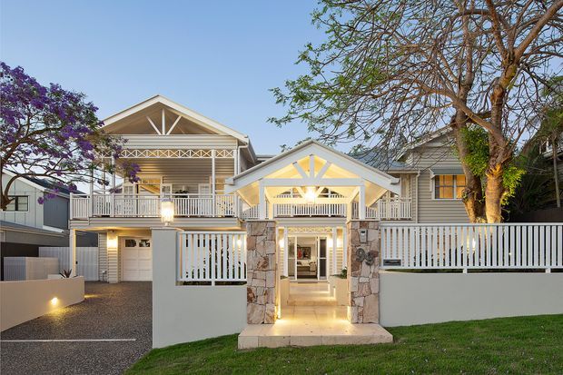 Front exterior view of a large two-storey residence with multiple pitched gables, white-painted verandah railings, timber weatherboard cladding, and a prominent covered entry canopy supported by stone-clad posts. The facade combines pale grey and white finishes with warm exterior lighting, landscaped lawn, mature trees, and a gravel driveway.