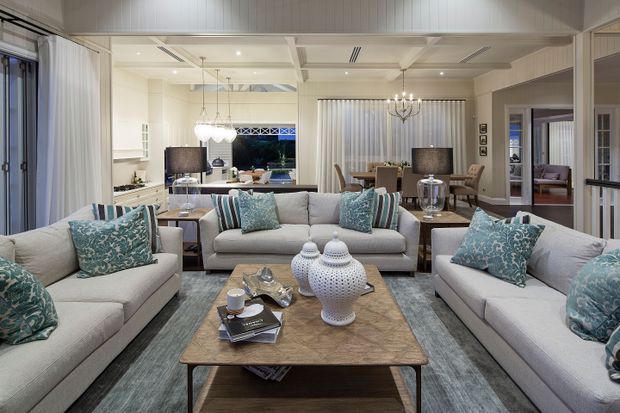 A bright, open-plan living room with three light upholstered sofas arranged around a square timber coffee table. Blue patterned cushions, grey-blue rugs, white ceramic accessories, sheer curtains, and a formal dining area create a refined coastal-inspired scheme. White panelled walls and ceiling beams, large glazed openings, recessed lighting, and a decorative chandelier are visible, with the kitchen and dining spaces extending behind the seating area.