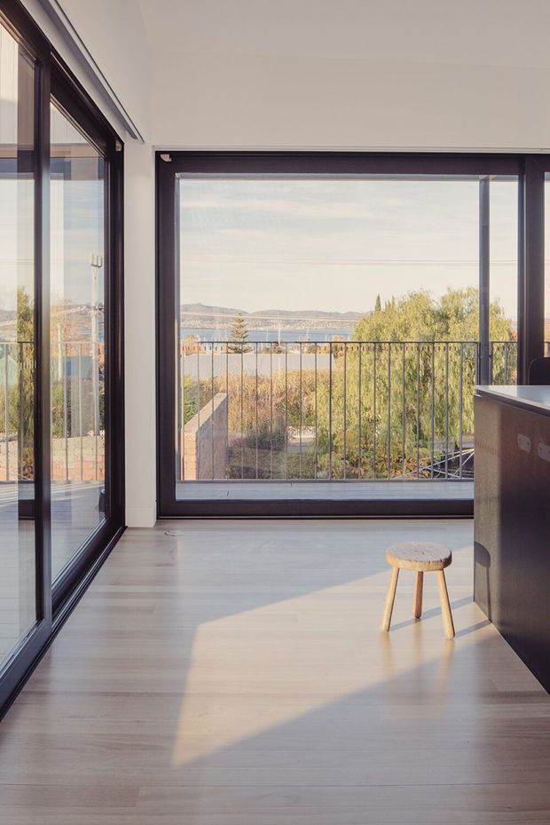 Bright, minimalist interior corner with expansive dark-framed glazing overlooking a balcony, trees, water and distant hills. Pale timber flooring, white walls and ceiling, and a dark kitchen island or counter create a clean, contemporary composition. A simple wooden stool sits in a strong patch of sunlight.