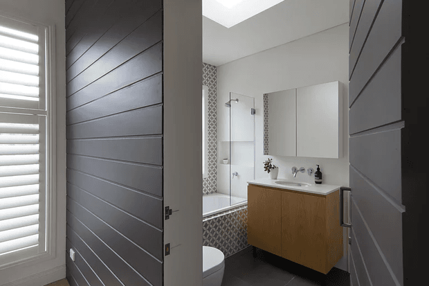 A contemporary bathroom viewed through a partially open horizontal-panel door. The room features a floating timber vanity with a pale benchtop, wall-mounted double mirrors, a glass-enclosed shower over a bathtub, patterned geometric wall tiles, dark floor tiles, and a skylight. A shuttered window and matching dark panelled doors frame the view.