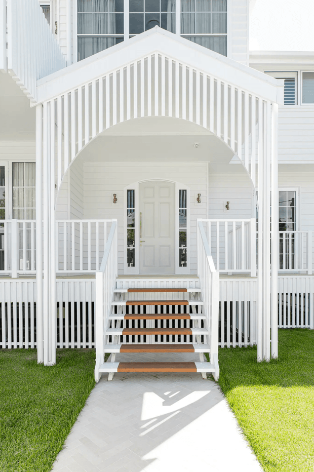 Front exterior entrance of a bright white timber-clad house, featuring a prominent gabled porch with decorative vertical balustrading, an arched entry canopy, white railings, timber-tread steps, and a paved path through a manicured lawn. Tall multi-pane windows and horizontal weatherboard siding reinforce the crisp coastal character.