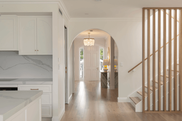Bright, refined residential interior showing a white kitchen at left opening to a timber-floored entry hall. A curved archway frames a paneled front door and chandelier, while a contemporary open timber-slat stair balustrade defines the right side. The space features white cabinetry, veined pale stone surfaces, detailed wall mouldings, and warm natural timber flooring.